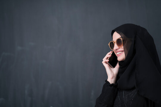 Young modern muslim businesswoman using smartphone wearing sunglasses and hijab clothes in front of black chalkboard