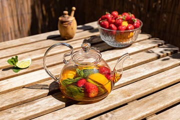 Fruit red tea with orange, strawberries, cinnamon, and fresh mint in a glass teapot on the sunny balcony. Trendy hard shadows.