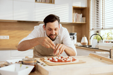 Handsome father dressing homemade pizza on wooden surface with feta cheese. Cooking baking preparing domestic pizza for birthday celebration making surprise for daughter son.