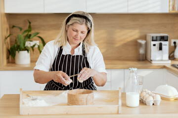 Middle-aged mother spending time in modern kitchen breaking egg pouring on flour preparing homemade...