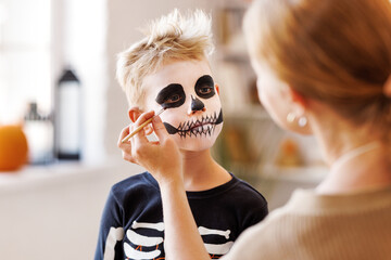 Festive makeup for Halloween. Woman doing skeleton make-up for boy in costume while preparing...