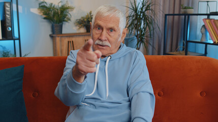 Hey you. Senior old man smiling excitedly and pointing to camera, choosing lucky winner, indicating to awesome you, inviting, approve. Elderly mature grandfather at home living room sitting on couch