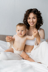 Happy curly mother holding hands of infant daughter on bed.