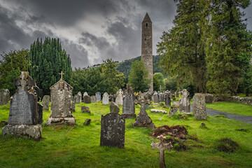 Medieval tower and ancient graves with Celtic crosses in Glendalough Cemetery. Moody storm sky in...