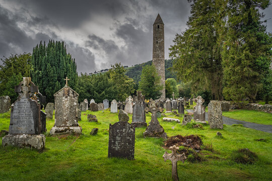 Medieval tower and ancient graves with Celtic crosses in Glendalough Cemetery. Moody storm sky in background, Wicklow, Ireland
