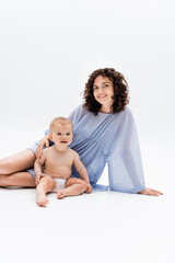 Happy woman in blue dress looking at camera near infant kid sitting on white background.