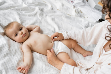 High angle view of mother wearing diaper on baby daughter on bed. - obrazy, fototapety, plakaty