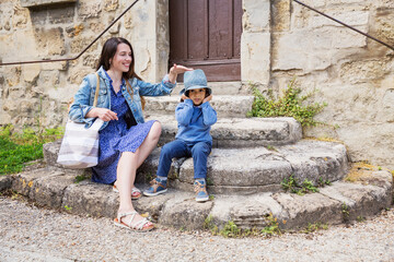 Fototapeta premium Mother and little handsome baby boy sitting on ancient stone stairs and playing outdoor with straw hat in old town