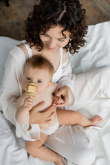 Overhead view of woman in pajamas holding infant daughter with toy on bed.