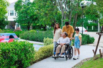 Asian elderly woman sitting on wheelchair which strolling by her happy family when walking at green...