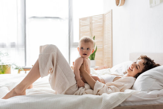happy woman in loungewear lying on bed and holding baby daughter.