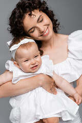 Joyful woman hugging smiling baby daughter in dress isolated on grey.