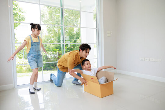 Happy asian father playing with cute small kids daughters and son laughing on moving day, boy having fun riding in box in new house.