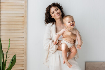 Brunette woman smiling at camera while holding baby girl near folding screen at home.