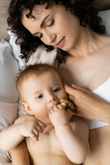 Top view of baby girl holding toy near mom on bed in morning.