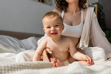 mother in loungewear with crop top sitting near happy infant daughter in bedroom.