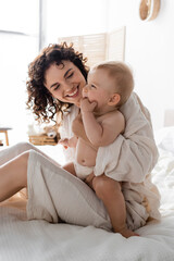 cheerful woman in loungewear holding in arms baby daughter and sitting on bed.