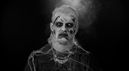 Sinister man with horrible scary Halloween zombie makeup in costume making faces, looking ominous at camera, trying to scare. Dead guy with wounded bloody scars face isolated against black background