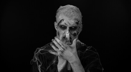 Zombie man with makeup with fake wounds scars and white contact lenses spells conjures over a candle, trying to scare in dark studio room. Sinister dead guy. Halloween, filming, staging concept