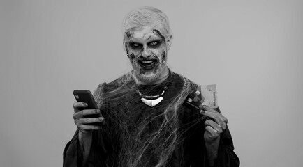 Sinister man with horrible scary Halloween zombie make-up using credit bank cards and mobile phone, transferring money purchases online shopping. Dead guy with wounded bloody scars face, gray room