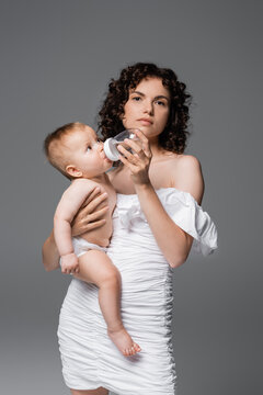 Trendy woman in white dress holding daughter and baby bottle while looking at camera isolated on grey.