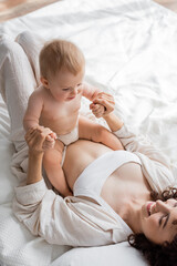 top view of happy woman in loungewear lying on bed and holding hands of infant daughter.