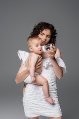 Stylish mom holding baby bottle and infant daughter isolated on grey.