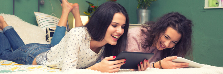 Two smiling girlfriends lying in bed and using smartphone and tablet 