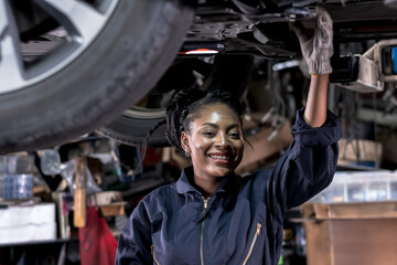 Obraz na płótnie Canvas Smiling African black car mechanic woman working underneath car in auto repair shop, Car maintenance concept