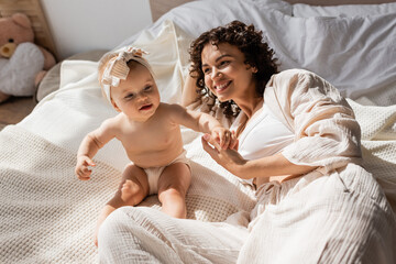 Fototapeta na wymiar happy mother with curly hair lying on bed and looking at infant daughter in headband.