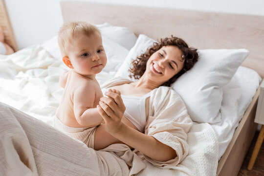 happy woman in loungewear lying on bed and holding hand of infant daughter.