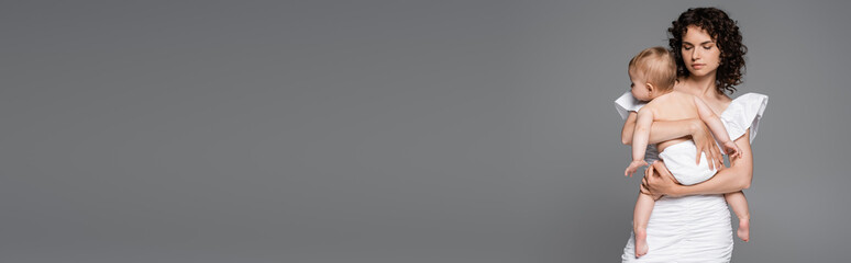 Woman in white dress hugging baby girl in panties isolated on grey, banner.