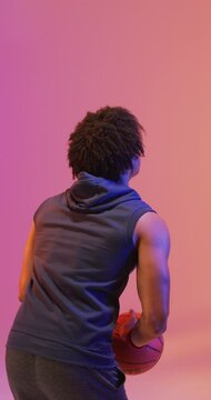 Vertical video of biracial male basketball player throwing ball on pink background