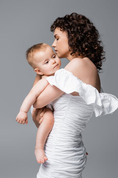 Curly woman in dress hugging infant daughter isolated on grey.