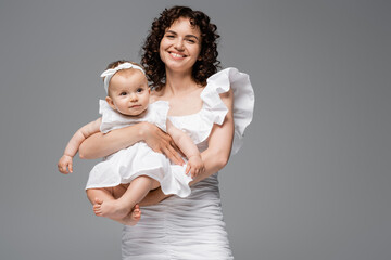 Smiling mother in stylish white dress holding baby girl isolated on grey.