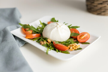 Fresh burrata cheese with arugula leaves, cherry tomatoes and walnuts on a white serving plate on a white table. Traditional Italian cheese
