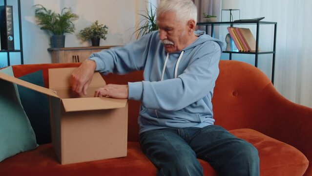 Happy senior grandfather unpacking delivery parcel sitting at home. Smiling satisfied elderly man shopper, online shop customer opening cardboard box receiving purchase gift by fast postal shipping