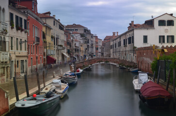 Long exposure of a Venetian street with boats on a canal in the foreground and a bridge in the background. People in motion are on the pavement.