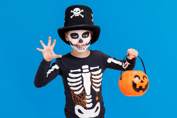 Happy cheerful boy in skeleton costume with  pumpkin  basket celebrates Halloween and scary gesture  on blue background