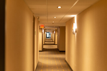 A long narrow hallway of a hotel with a red exit sign light sconce lights and cream colored walls. The carpet on the floor is green and beige with a pattern. The low ceiling has white tiles. 
