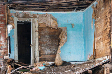 The interior of a building being demolished. The walls are layers of blue paint, wallpaper, and wood. There's an open single white wooden door in the house. The room is covered in dirt and materials. 