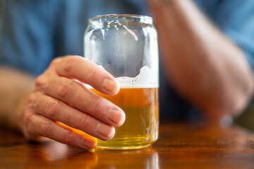 A male reaches for a tall clear glass of sour craft beer in a microbrewery. The cold refreshment has frost on the glass. The beer is on a table and coaster. The top of the glass has white froth. 