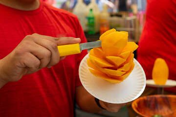 Mexican mango flower or mango on a stick being made by a street vendor at a market. The fruit is orange color and the vendor is sprinkling lime, chili, salt, and cinnamon over the sticky sweet fruit. 