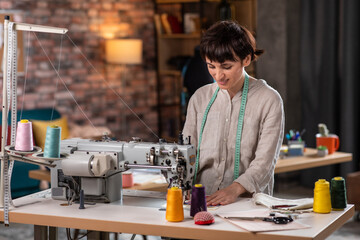 Fototapeta premium Beautiful young woman dressmaker very carefully sew some fabrics on the professional sewing machine she smiling pretty in a modern tailoring fashion atelier