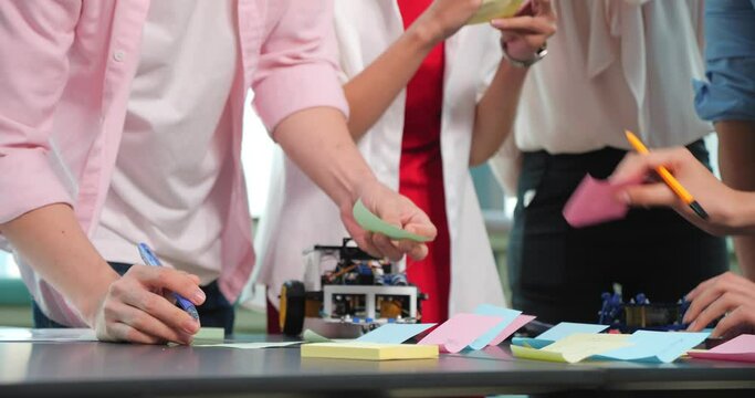 Group of high school students building and programming electric toys and robots in robotics classroom, brainstorm, exchange ideas, and writing on colorful sticky post-it notes.