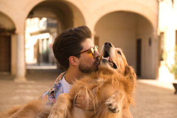 Portrait of young Hispanic man with beard and sunglasses kissing his dog which he is holding in his arms very happy. Concept animals, dogs, love, pets, golden.