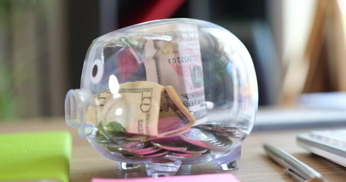 Coins and banknotes fall into transparent piggy bank