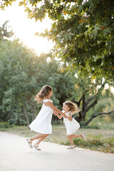 Naklejka na ściany i meble Two cute siblings girls wear similar white dress hoding hands and dance together in park over green nature background outdoor. Childhood. Friendship.