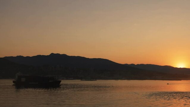 Sunrise just peeks over Vancouver Harbor with mountains and foreground ship in silhouette in the golden hour at first light of day.