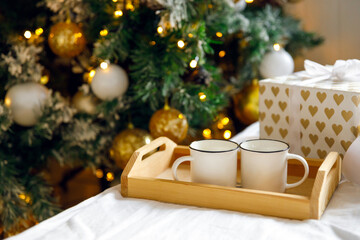 Gift box, wooden tray and two white mugs on the bed on a white sheet, near a decorated Christmas tree. Cozy morning. Coffee in bed
